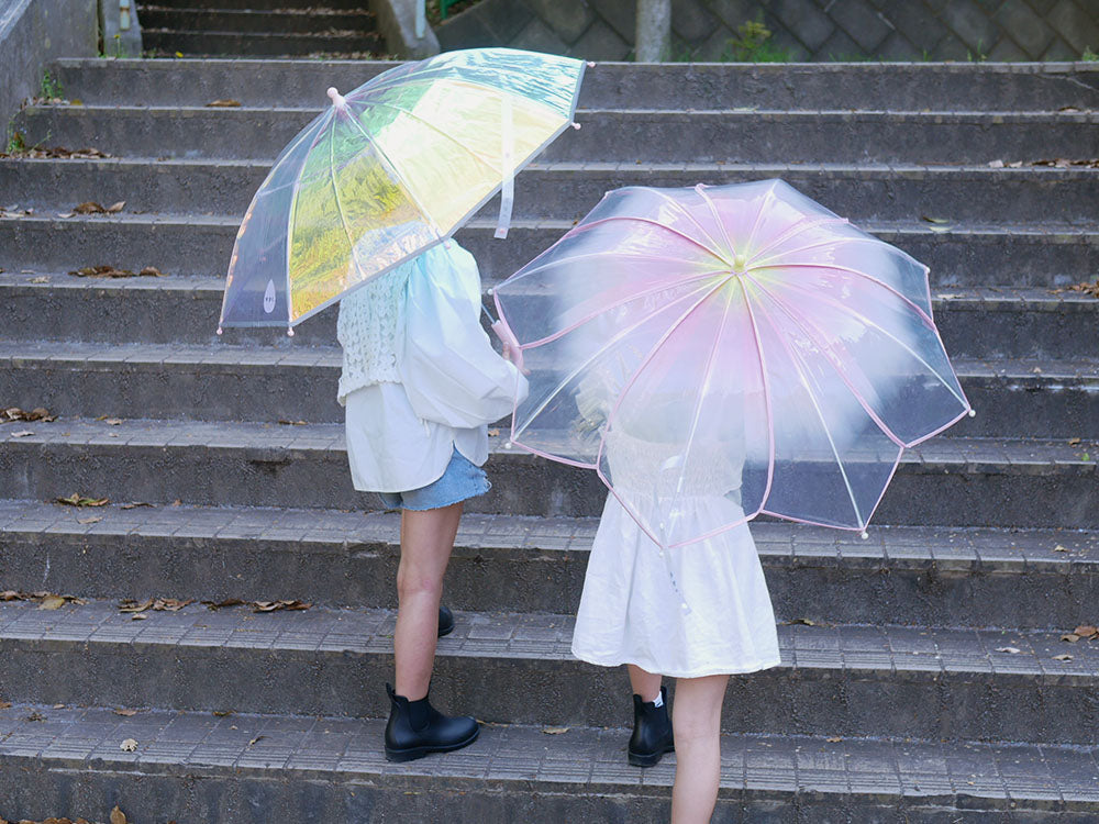 レインコートを着てピンクのキッズシャイニーアンブレラをさし、雨の中を歩く子供の姿