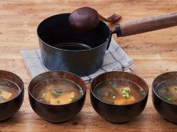 藤田金属の「今日を元気にするお味噌汁パン」に具だくさんの味噌汁が盛られ、キッチン用品と共にテーブルに置かれている様子