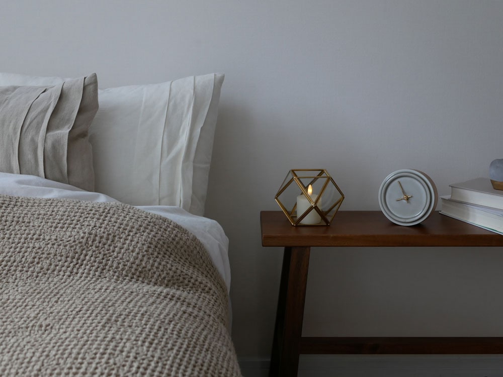 木製ベッドフレームの上に真鍮製ガラスケース内のフィギュアが配置され、リネンのベッドシーツとスローピローが整えられたベッドルームの様子