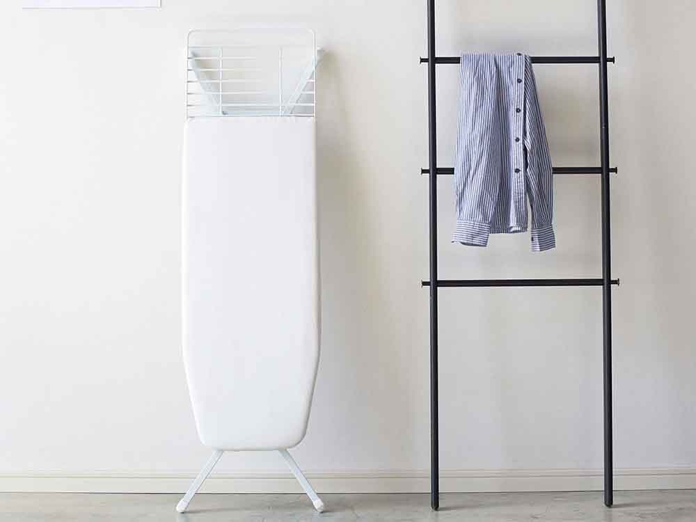 灰色のスタンド式アイロン台と衣類ハンガーが棚に配置された家庭用洗濯スペース