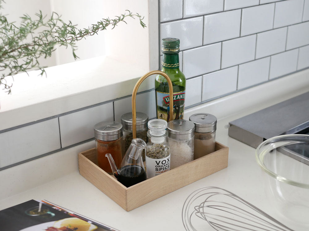 フリッツ・ハンセンのSkagerak Norr Carrierがオーク材と真鍮仕上げのキッチンカウンター上に配置され、食器や調理器具、果物、銀製ボトルが並べられている様子