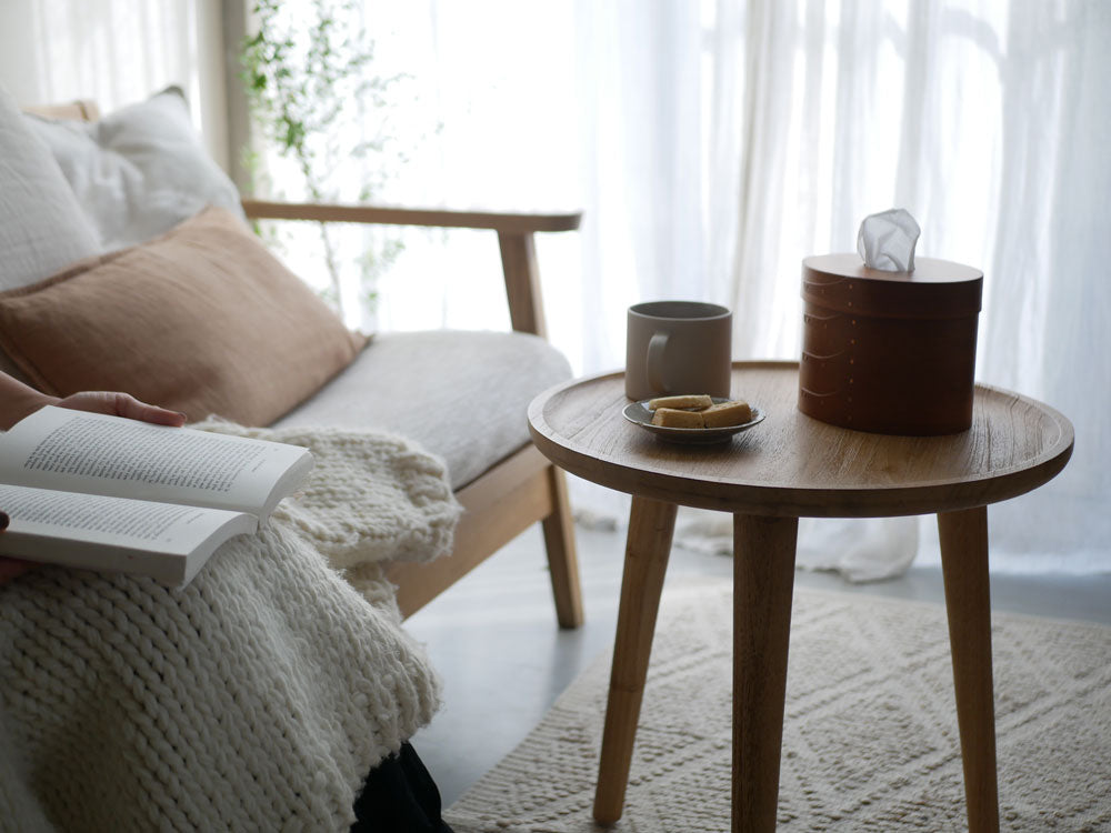 チェリー材のアクシス シェーカーティッシュボックスが木製コーヒーテーブルの上に置かれ、花鉢やビーズクッションとともにスタイリッシュに配置された室内インテリア