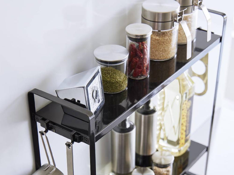 スチール製の2段コンロ横ラックがキッチンに設置され、上段にはテーブルウェアやグラスが、下段には調理器具や小型家電が整然と収納されている様子。