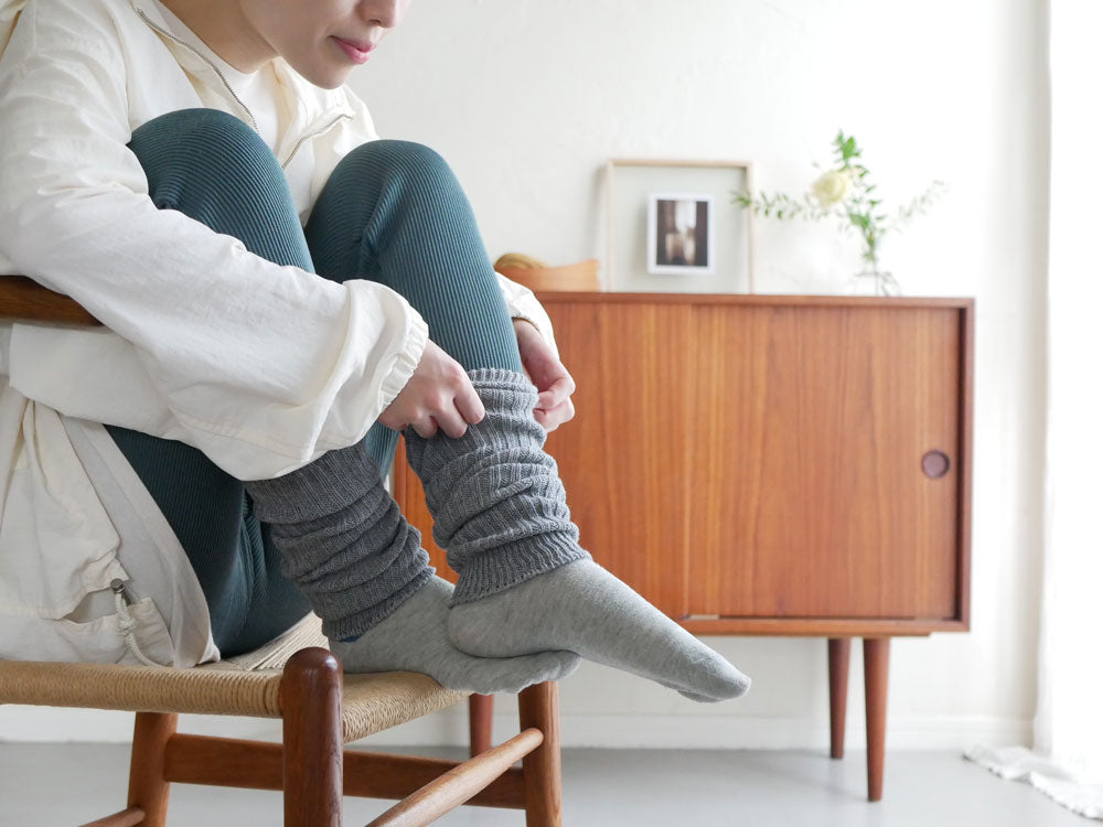ベージュ色のmemeriカシミヤコットンアーム＆レッグウォーマーを装着した裸足の脚が木製の床と家具の上に置かれ、快適な雰囲気を表現している