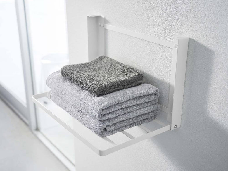 プラスチックハンドル付きスチール製タワー折り畳み棚がバスルームの石こうボード壁に設置され、タオルや日用品が収納されている様子