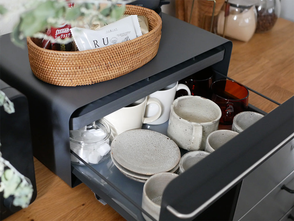 ピュアホワイトの【UtaU】カウンタードロワーがキッチンに設置され、スチール製の棚や引き出し、ステンレス取っ手と花鉢が配置されたモダンな収納家具