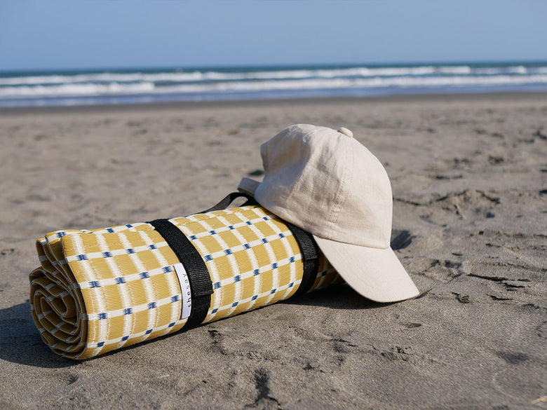 海辺の砂浜にシセイの洗えるレジャーマットが敷かれ、ビーチバッグが置かれたリラックスした風景