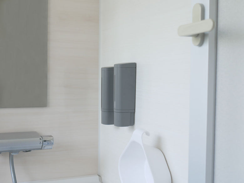 グレーの浴室壁に取り付けられたリッチェル下から出るシールフックディスペンサーと、隣接するハンドドライヤーやトイレットペーパーホルダー