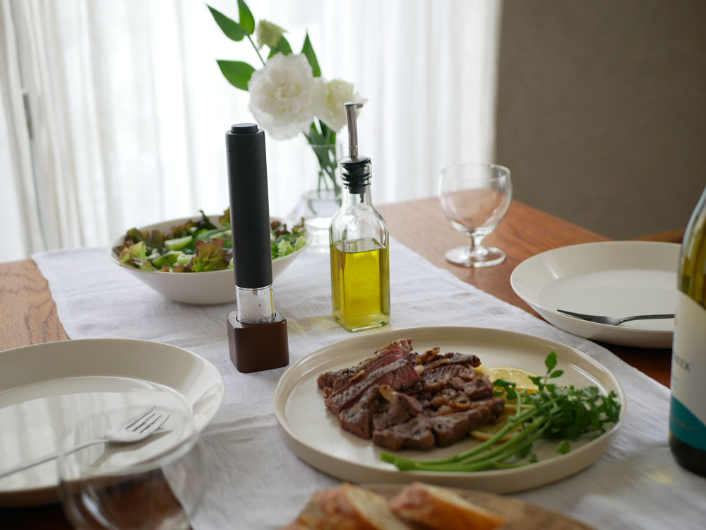 レコルトのアカシアスタンド付き電動ソルト＆ペッパーミルがテーブルに置かれ、食器や調味料とともにスタイリッシュに配置されています