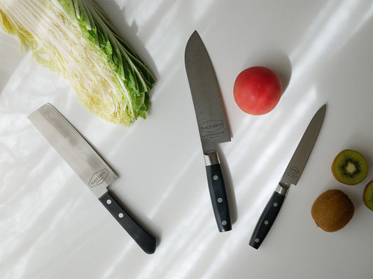 ダマスカス鋼TOOLSブラックスミスナイフシリーズのキッチンナイフが切り板と新鮮な野菜と共にキッチンに並べられている様子