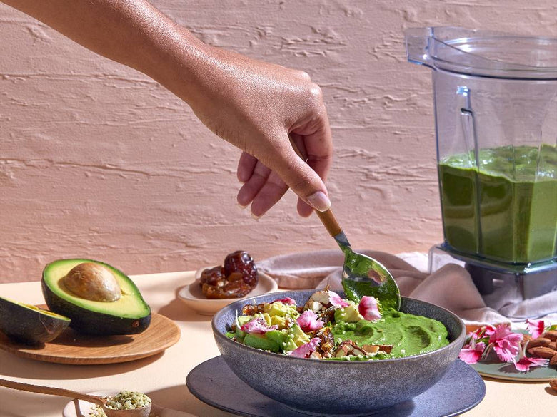 Vitamixブレンダーを中心に、テーブルに新鮮な野菜や果物、ボウルなどの食器が配置されたキッチンのシーン