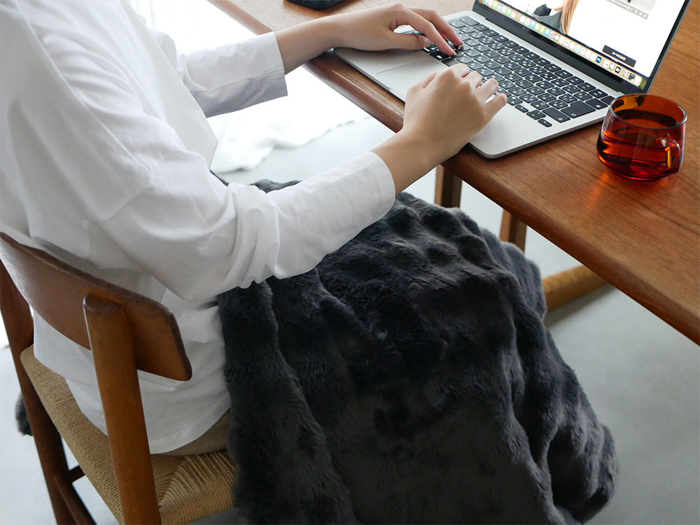 QUADSの掛敷両用電気毛布toronが木製のデスク上に広げられ、ラップトップや家具と共に配置され快適な作業空間を演出している様子