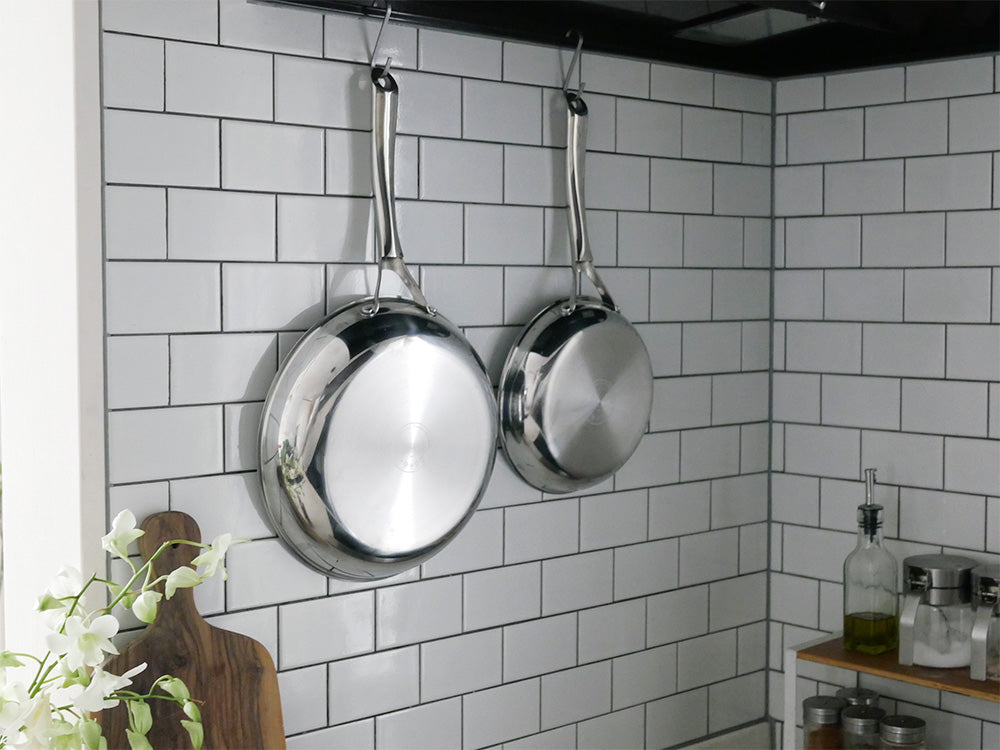 コレールのこびりつきにくい次世代フライパンと中華鍋がキッチンのポットラックに吊るされ、シルバーの調理器具がカウンターに整然と並ぶ様子