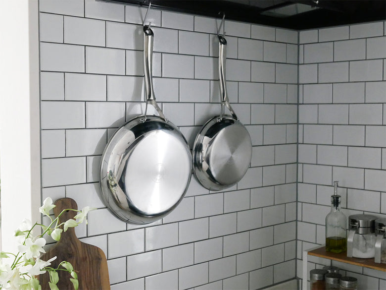 コレールのこびりつきにくい次世代フライパンと中華鍋がキッチンのポットラックに吊るされ、シルバーの調理器具がカウンターに整然と並ぶ様子