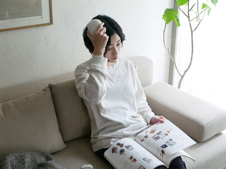 リビングルームのソファに座り、クッションに寄りかかりながらFESTINO充電式EMSヘッドスパを使用している様子